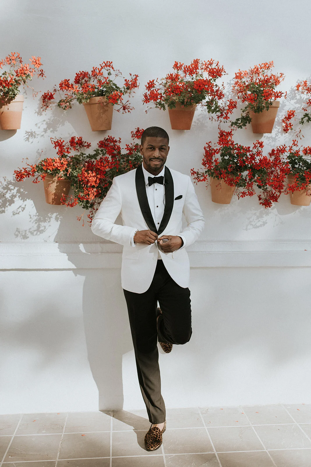 A man in a white tuxedo jacket with black lapels, black bow tie, and black pants stands against a white wall with red flowering plants in terracotta pots. He is smiling and holding the edge of his jacket with one hand, with one leg crossed over the o