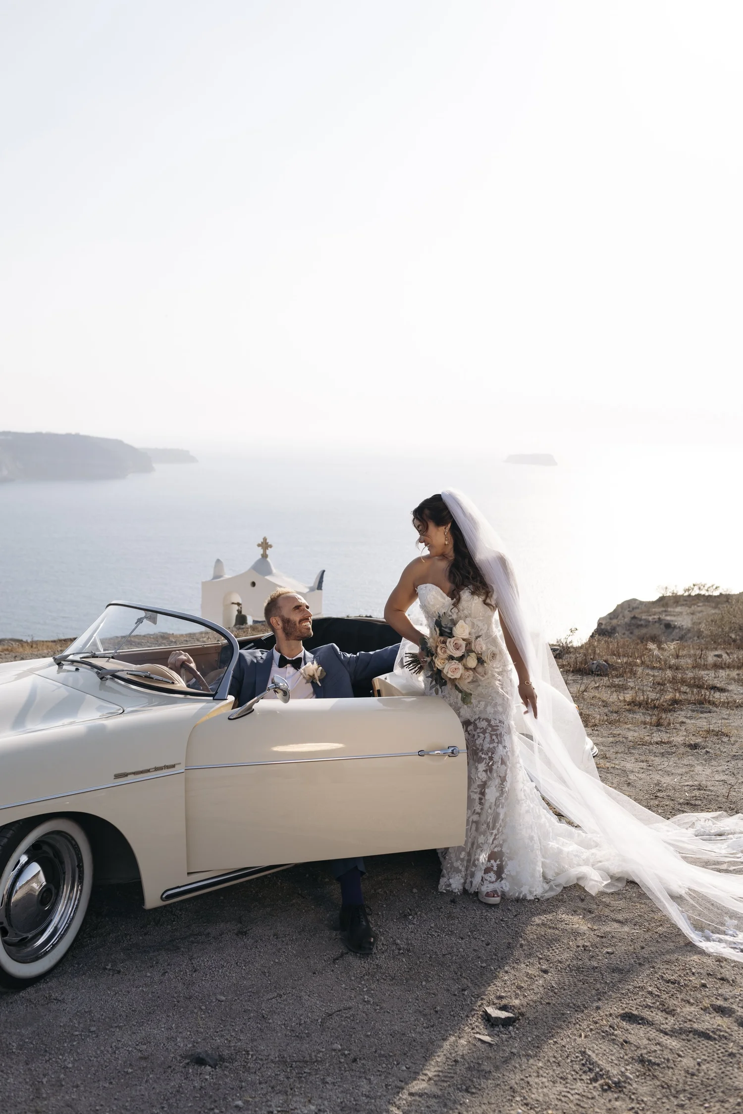 A bride and groom on a wedding day, with the groom sitting in a vintage convertible car and the bride standing next to him, holding a bouquet, in a scenic coastal setting with water and cliffs in the background.