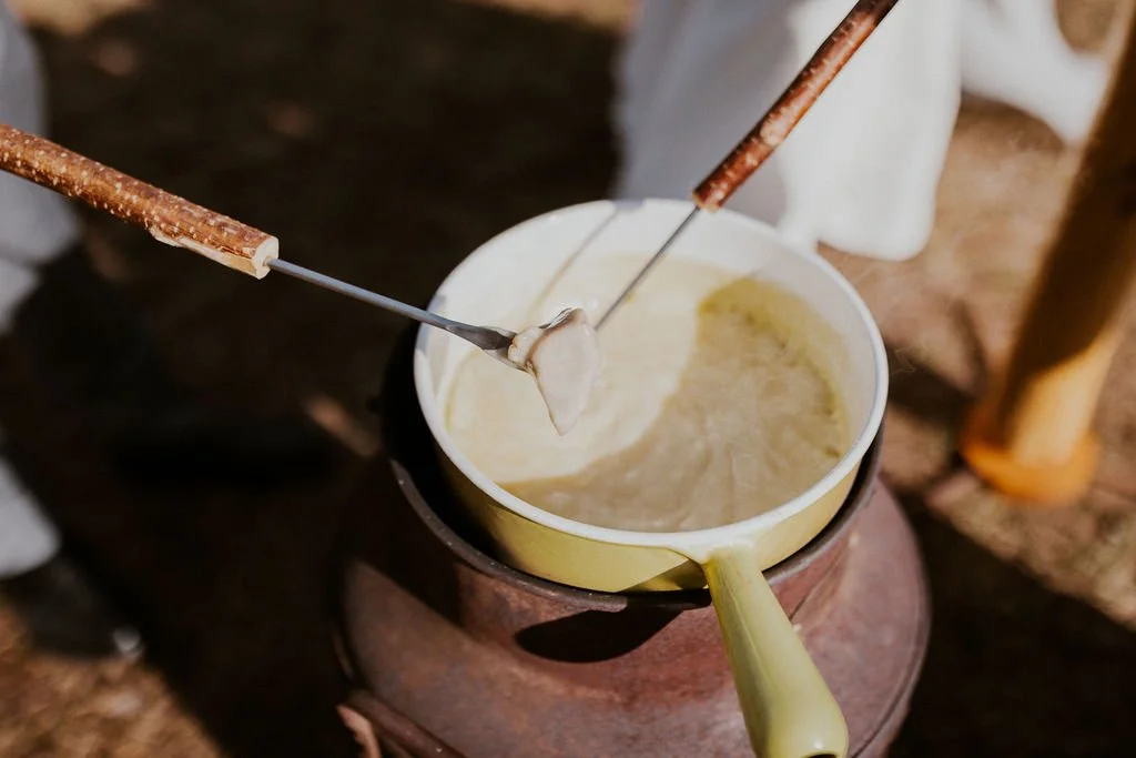 A mug filled with melted cheese with two skewers and a dollop of cheese on the skewers, placed on a wooden surface outdoors.