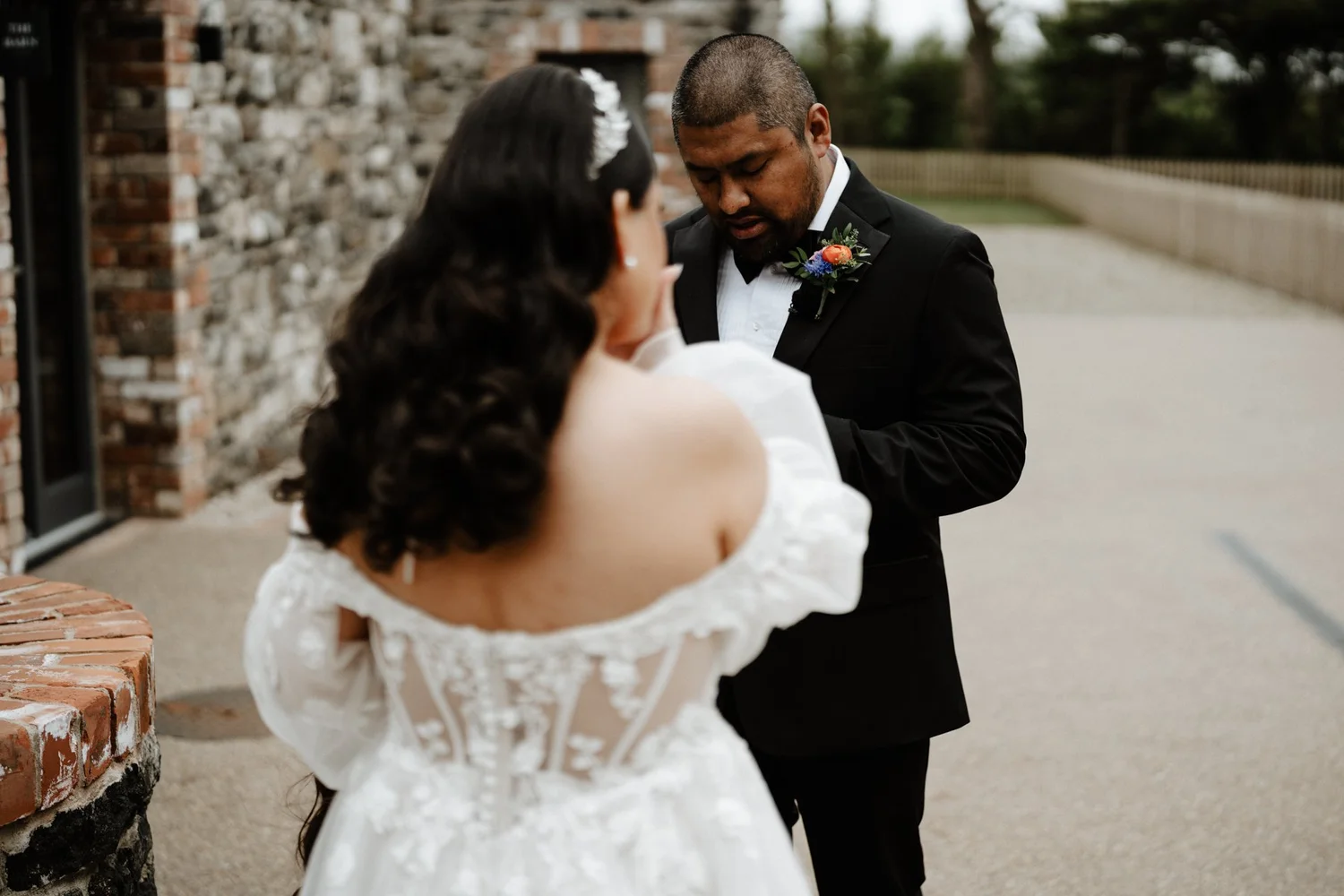 A couple dressed in wedding attire outdoors, with the woman in a white lace dress with open shoulders, and the man in a black tuxedo with a boutonniere, standing close and facing each other.