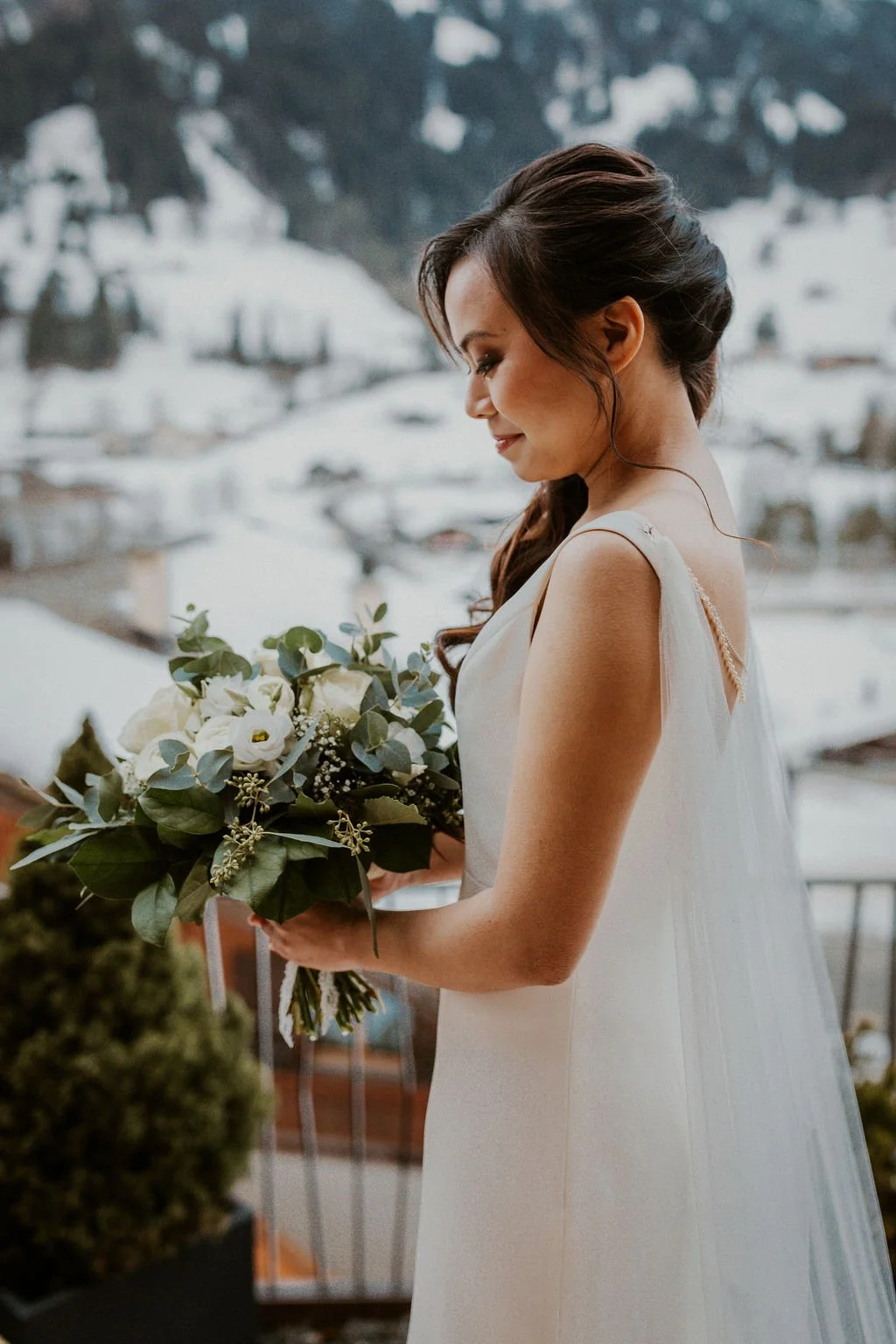 A woman in a wedding dress holding a bouquet of white flowers and greenery, standing outdoors with snow-capped mountains in the background.
