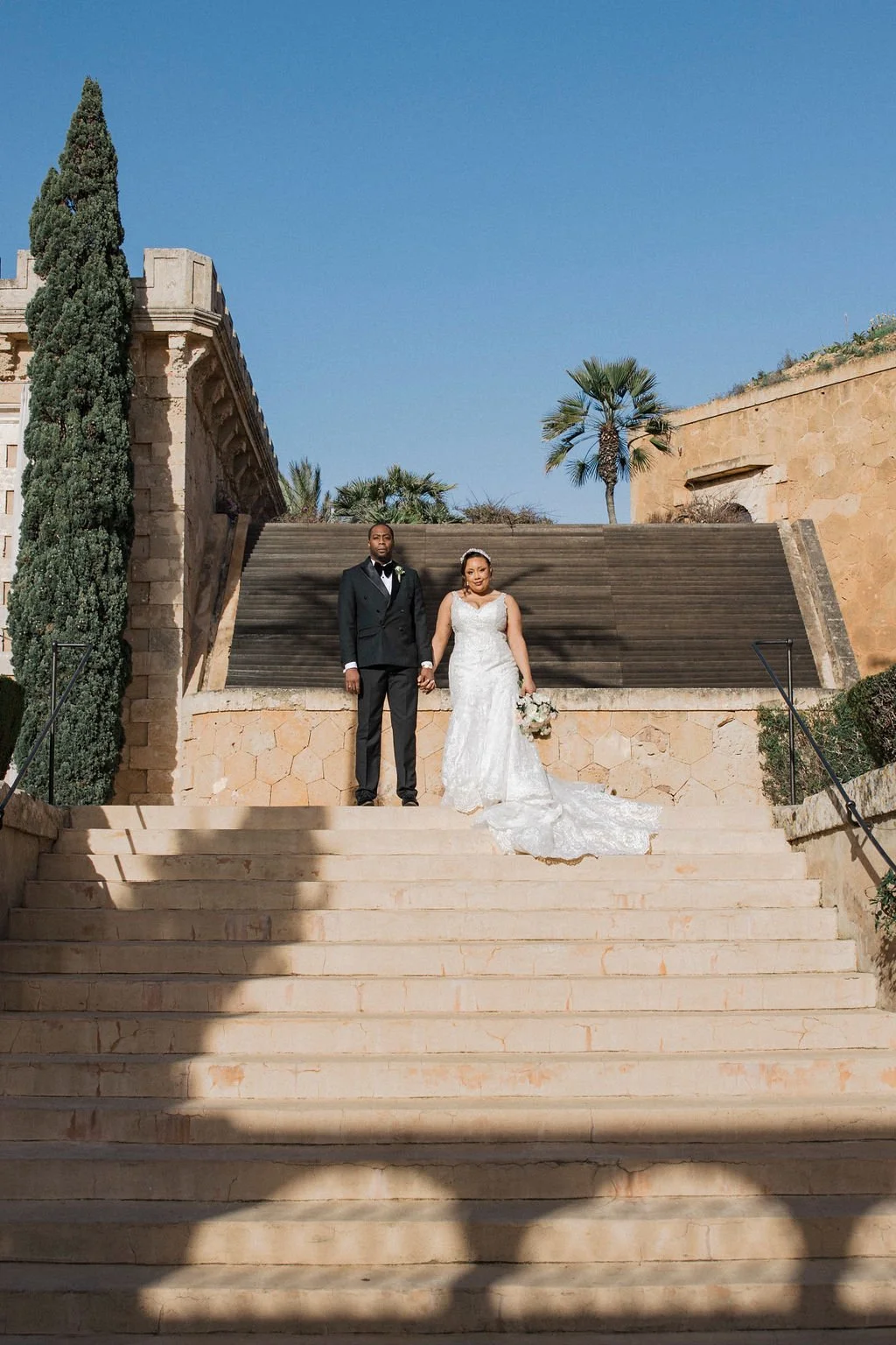 A bride and groom standing hand in hand on outdoor stairs, with bride in a white wedding gown and groom in a black tuxedo, against a backdrop of blue sky, palm trees, and historic stone architecture.