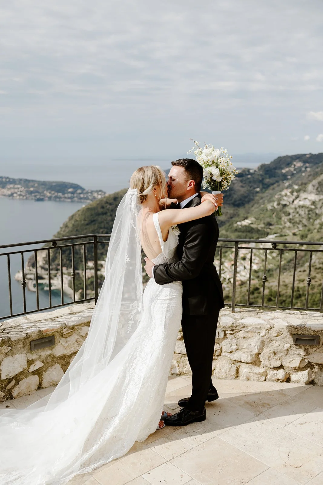 A bride and groom sharing a kiss on an outdoor terrace overlooking water and hills, with the bride holding a bouquet and wearing a white lace wedding dress with a long veil.