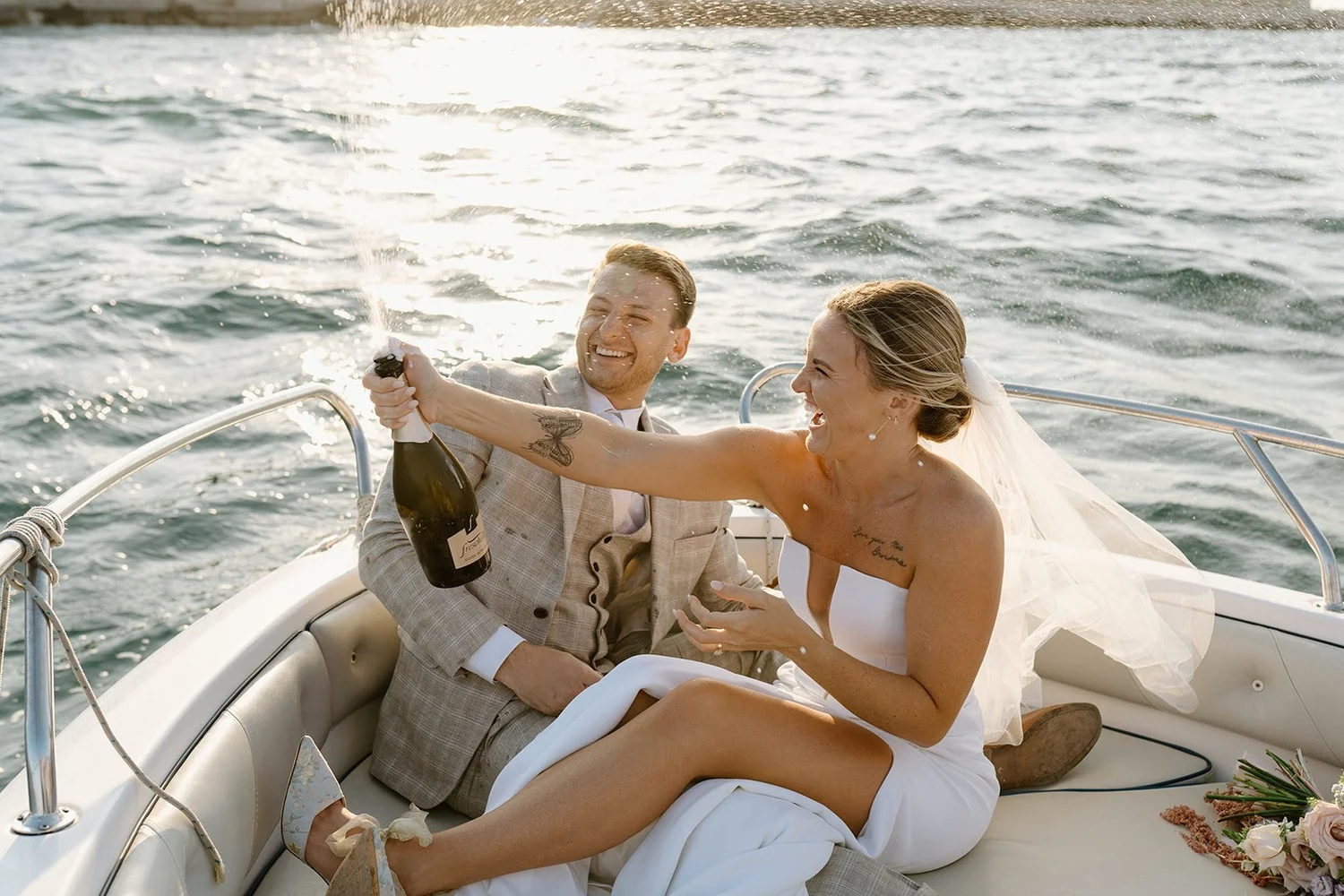 A bride and groom celebrating on a boat, with the bride spraying champagne and both of them smiling, water in the background.