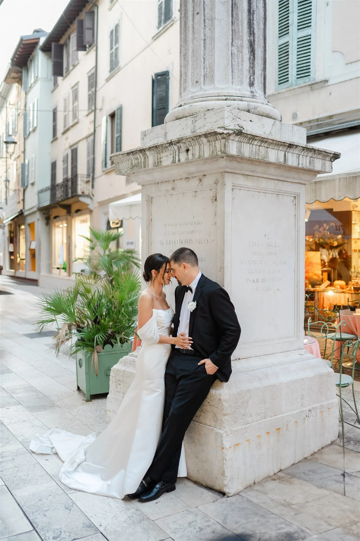A couple in wedding attire, a woman in a white wedding dress and a man in a black tuxedo, are standing close and touching foreheads in an outdoor urban setting near a white stone monument. The woman is holding the man's hand, and they appear emotional and intimate.