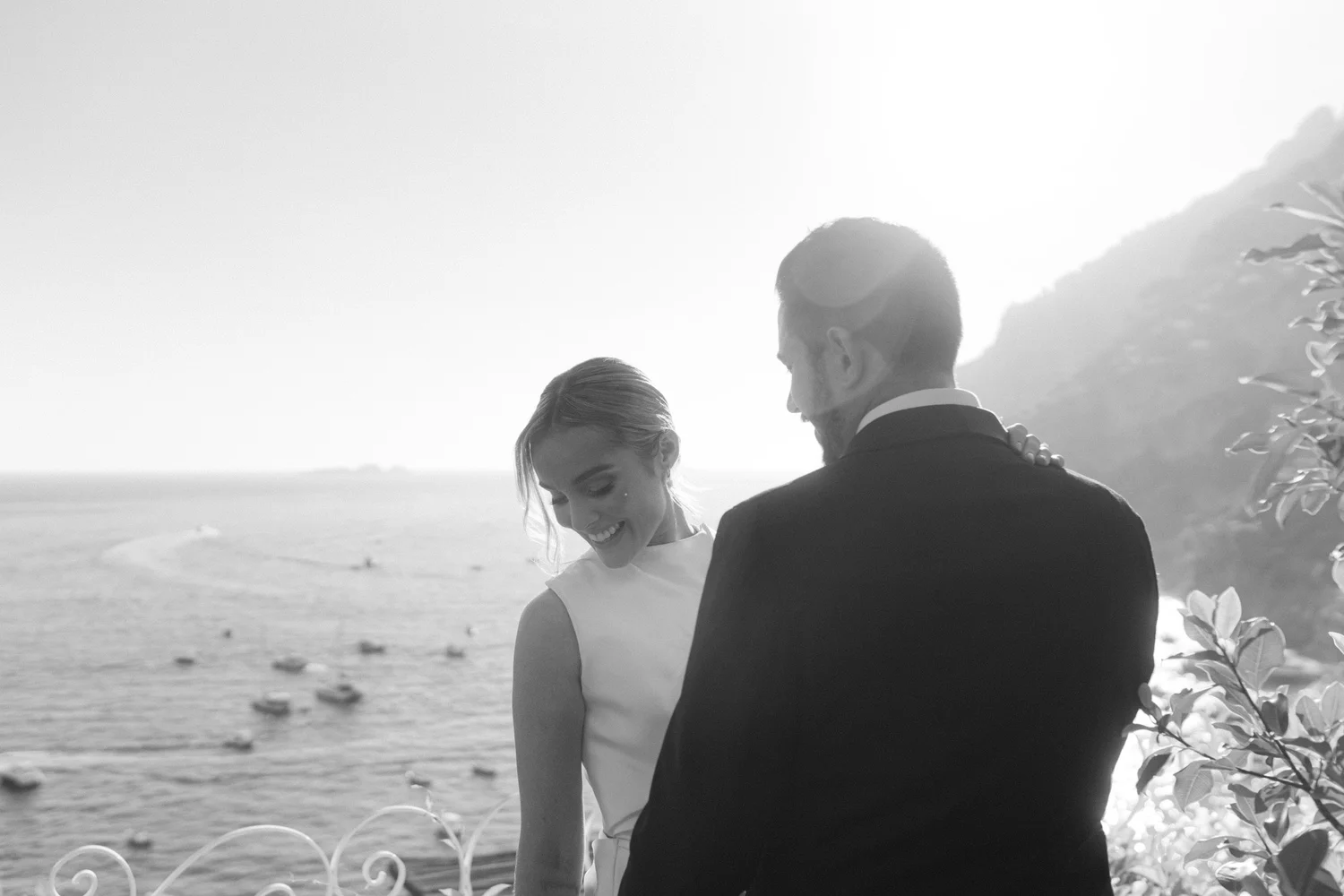 A black-and-white photograph of a joyful couple at a beach, smiling and embracing each other, with the ocean and the horizon in the background.