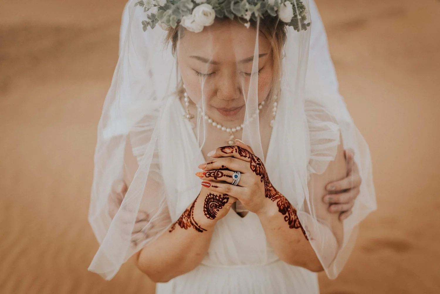 A woman dressed in white with a floral headpiece, wearing pearl jewelry, and showing intricate henna designs on her hands, with her eyes closed.