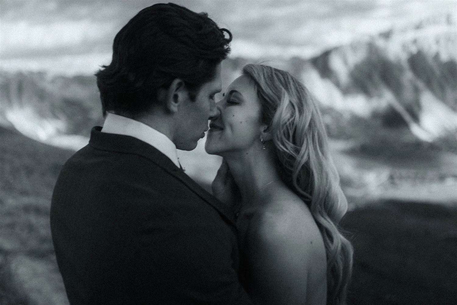 A black and white photo of a couple close together, about to kiss, with a mountain landscape in the background.