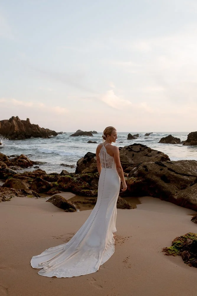 A woman in a white wedding dress standing on a sandy beach near rocks, with the ocean and sky in the background during sunset.