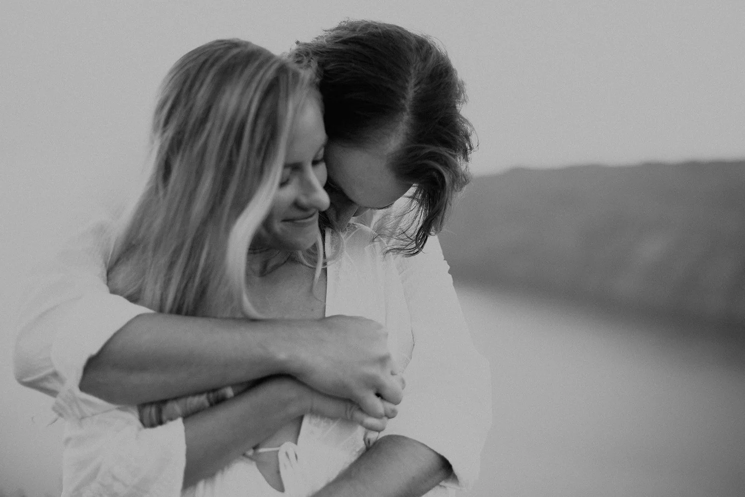Two women embracing each other with their heads bowed down and eyes closed, sharing a tender moment outdoors.