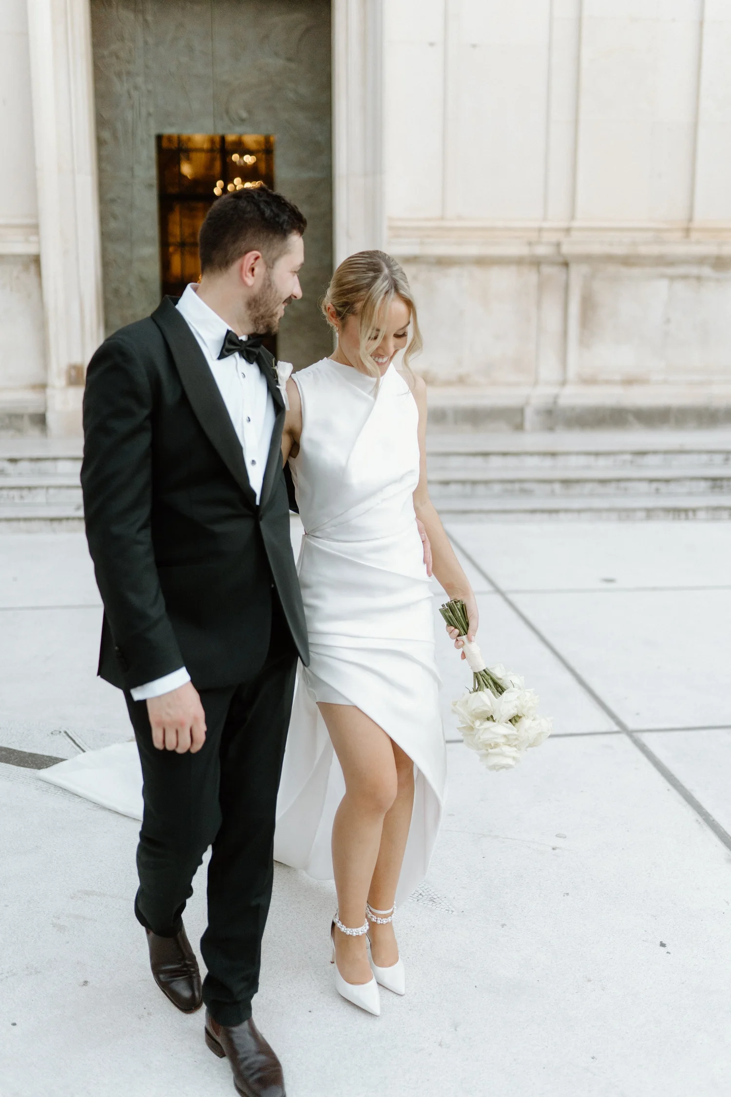 A bride and groom are walking together outside in front of a large stone building. The bride is wearing a white gown with a slit, white high heel shoes, and pearl ankle bracelets, and is holding a bouquet of white roses. The groom is dressed in a bla
