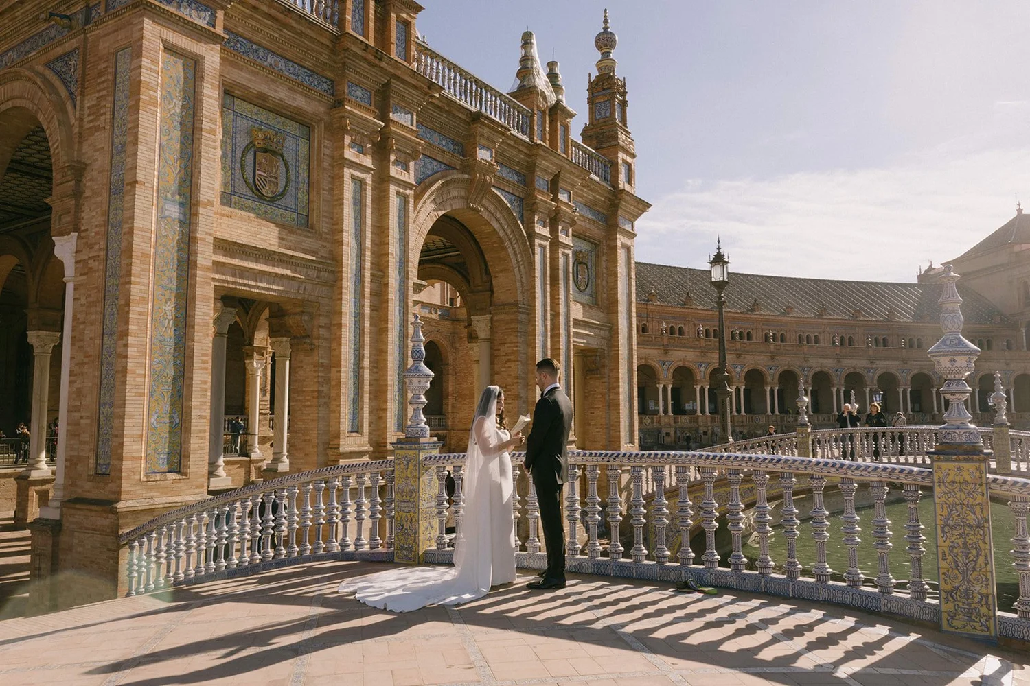A bride and groom stand on a ornate terrace with intricate tile and stonework, exchanging vows during their wedding ceremony outside a historic building with detailed architecture.