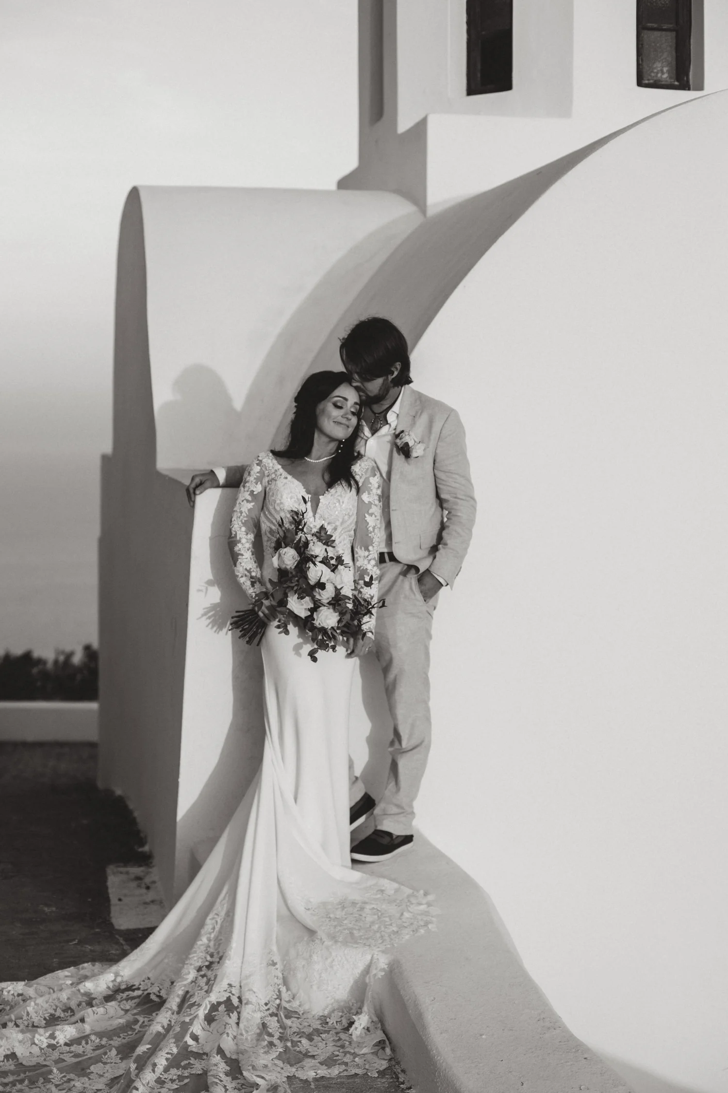 Black and white photo of a bride and groom standing together outdoors, with a white architectural structure and clear sky in the background. The bride is holding a bouquet of flowers, and both are smiling.