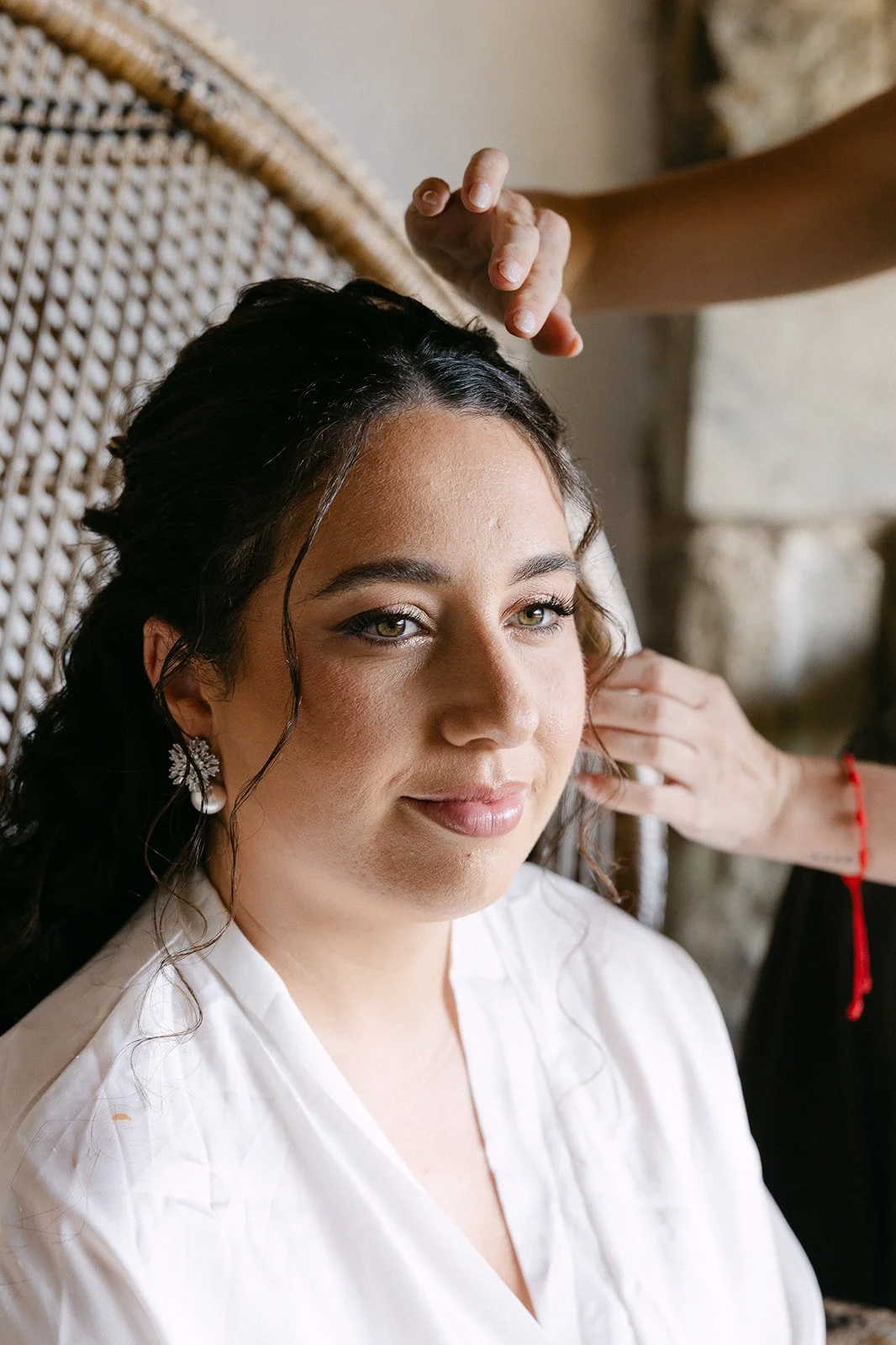A woman with dark, wavy hair and green eyes sits calmly while a person styles her hair.