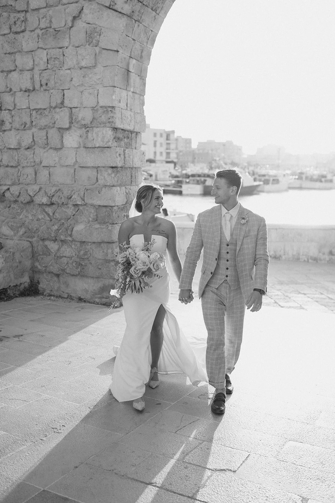 A black-and-white photo of a bride and groom walking hand-in-hand by a waterfront, smiling at each other, with boats and buildings in the background.