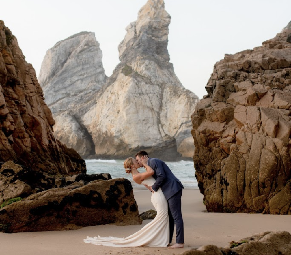 A couple in wedding attire sharing a kiss on a sandy beach surrounded by large rock formations with a mountain backdrop.