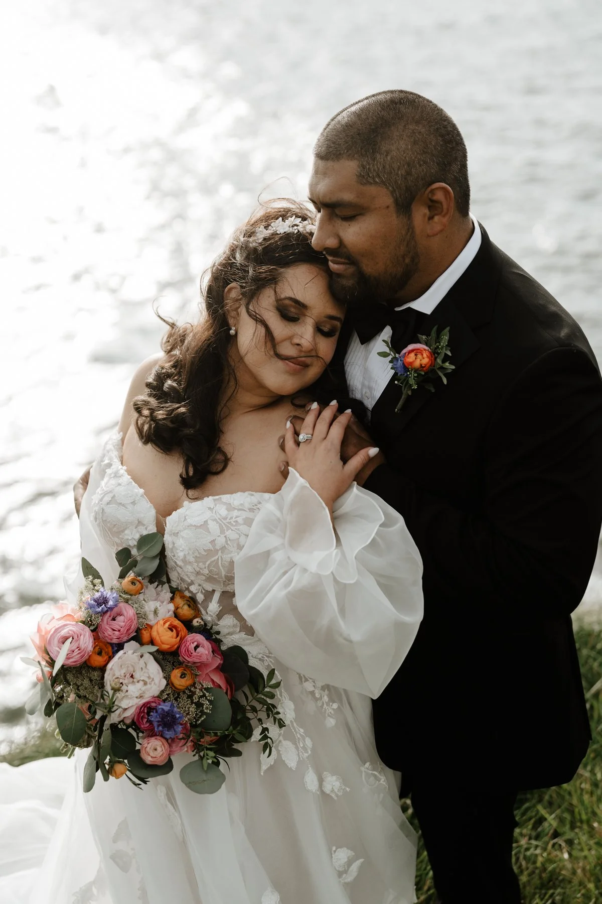 A bride and groom embrace by the water. The bride wears a white wedding gown and holds a colorful bouquet, while the groom wears a black tuxedo with a boutonniere. They appear to be peacefully celebrating their wedding.