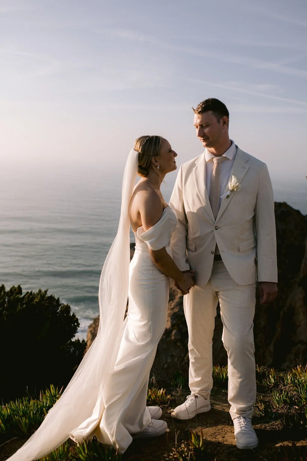 A bride and groom standing on a rocky cliff overlooking the ocean at sunset, holding hands and gazing into each other's eyes in Portugal.