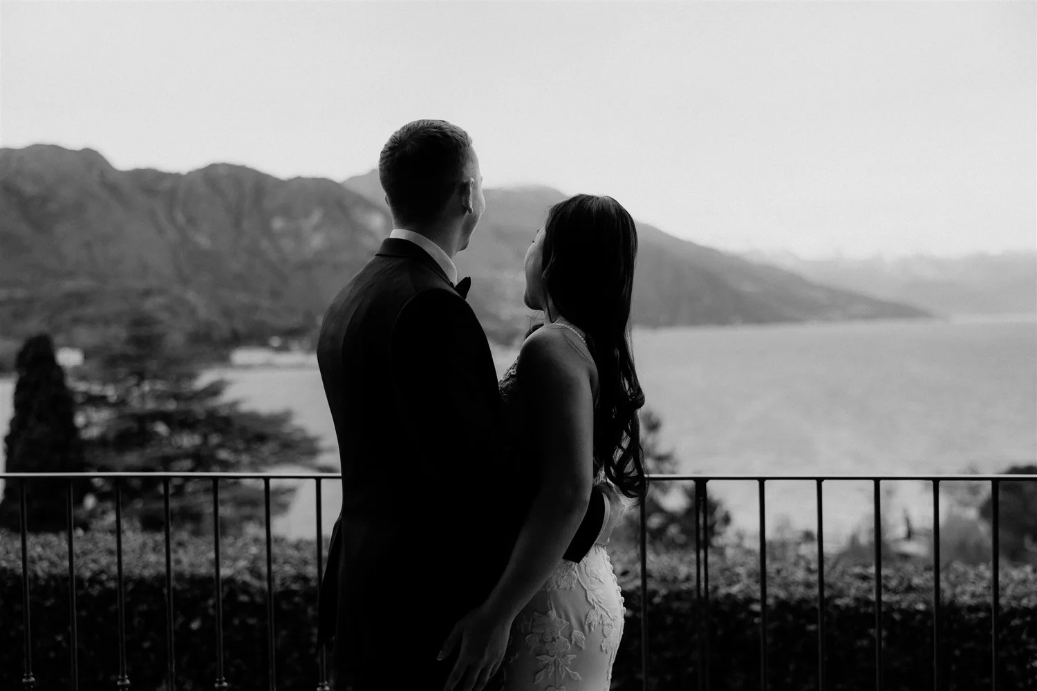 A black and white photo of a man and woman standing close together, gazing at a mountainous landscape over a body of water, with a railing in the foreground.