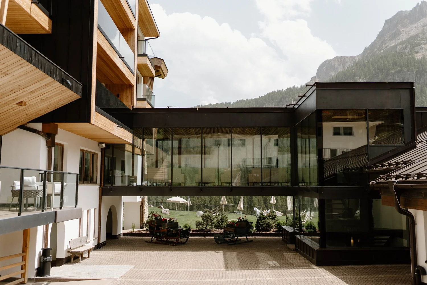 Modern building with wooden and black metal accents, glass walls, and outdoor seating area with umbrellas, mountain scenery in the background.