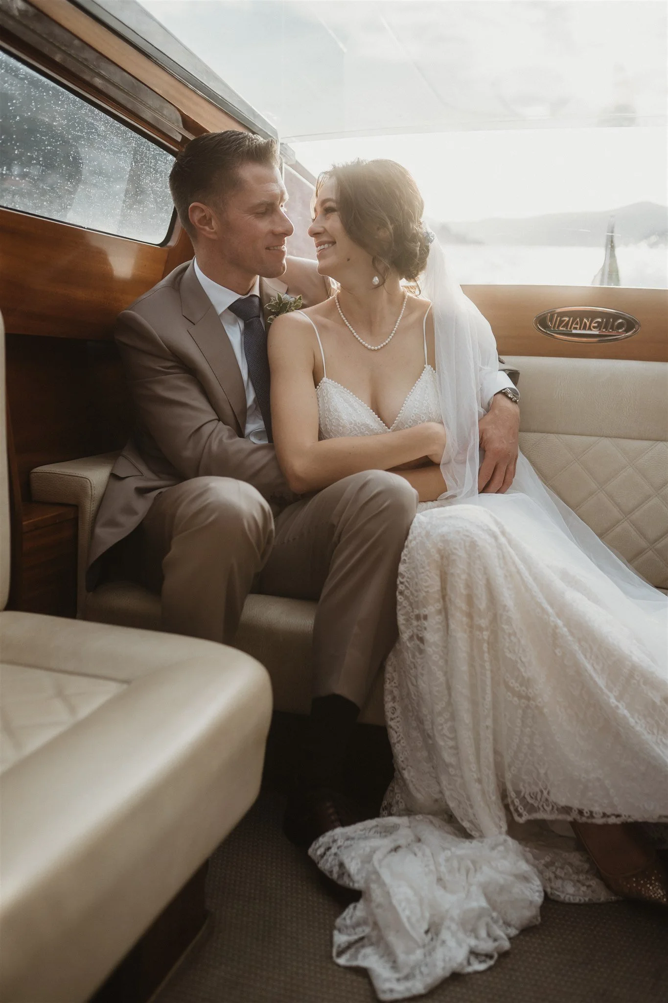 A bride and groom sitting closely inside a vintage boat, sharing a joyful moment, with the bride wearing a lace wedding dress and pearl necklace, and the groom in a beige suit with a boutonniere.
