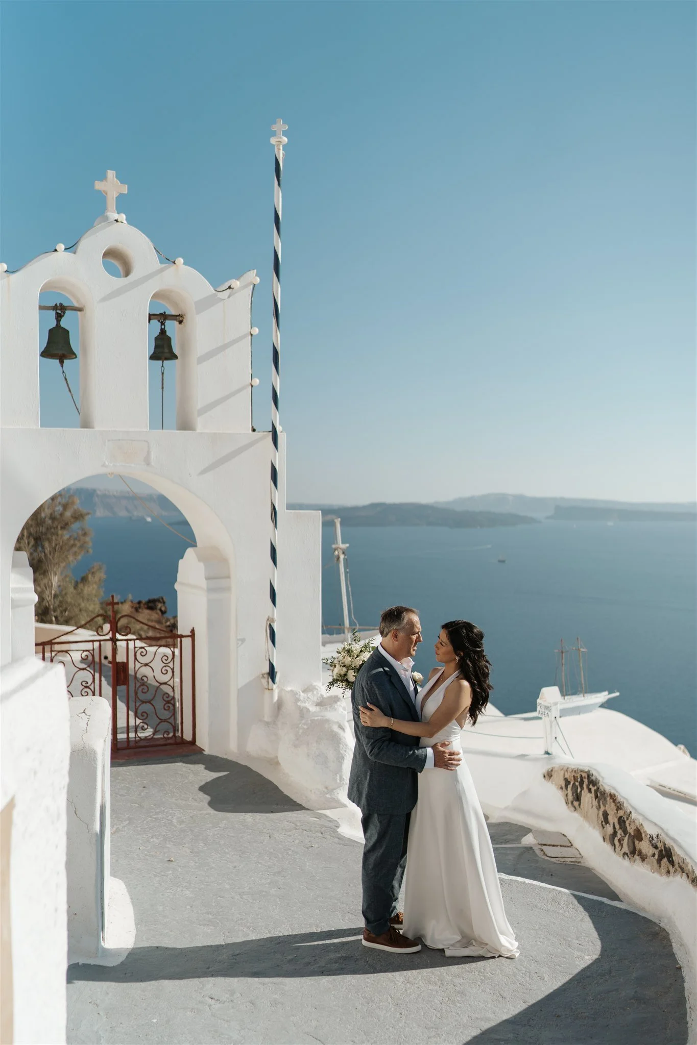 A bride and groom standing closely together in front of a seaside white chapel, with blue sky and ocean in the background, during a wedding ceremony.