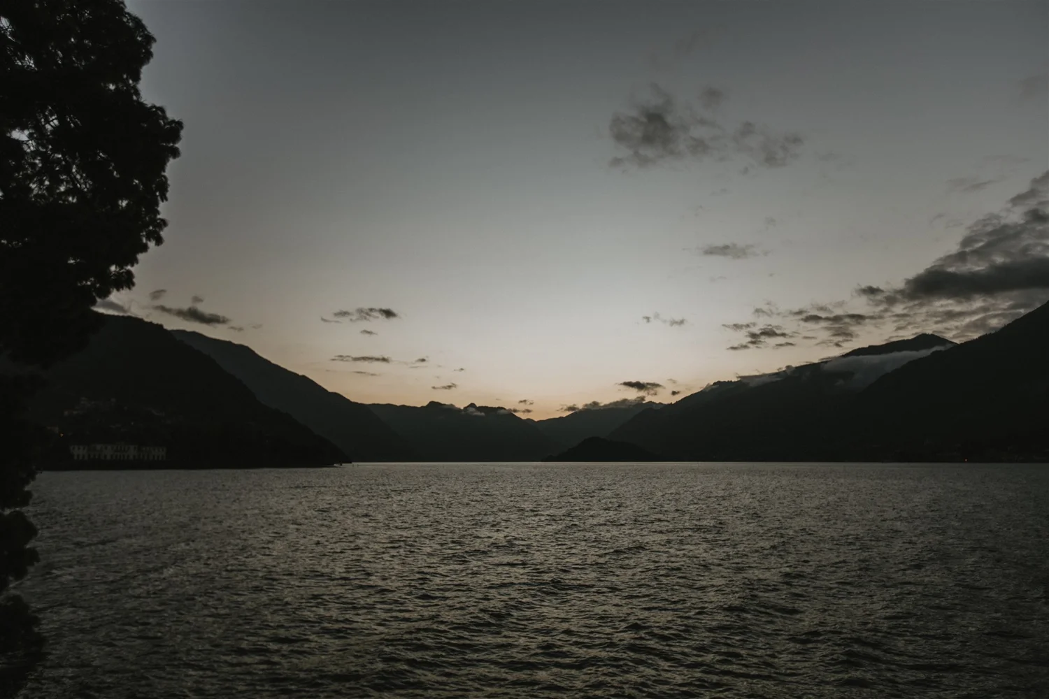 Serene lake surrounded by mountains during dusk, with a large tree on the left and a few clouds in the sky.