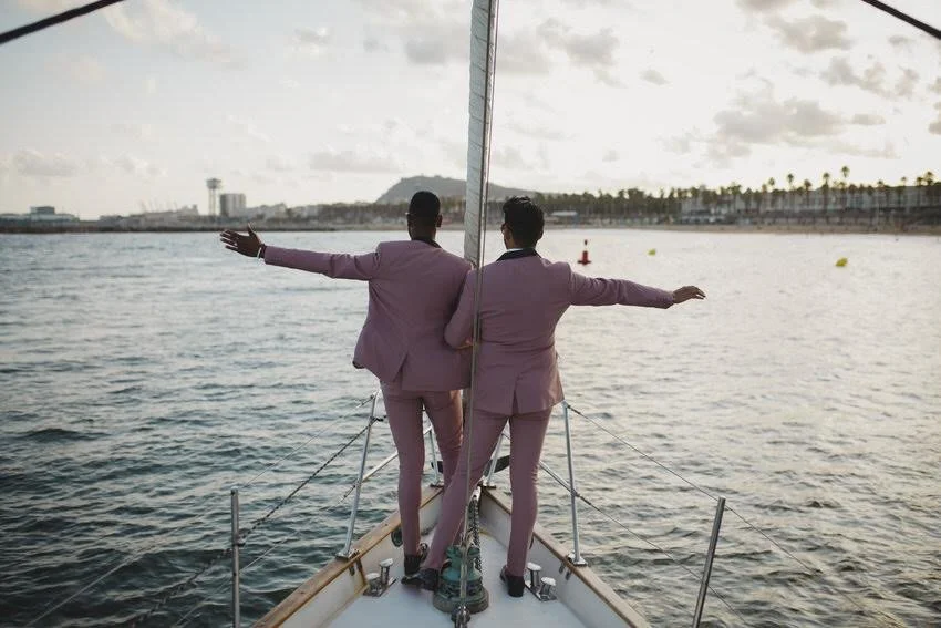 Two men in pink suits standing on a boat facing away, with arms outstretched, near a body of water and a distant shoreline with palm trees.