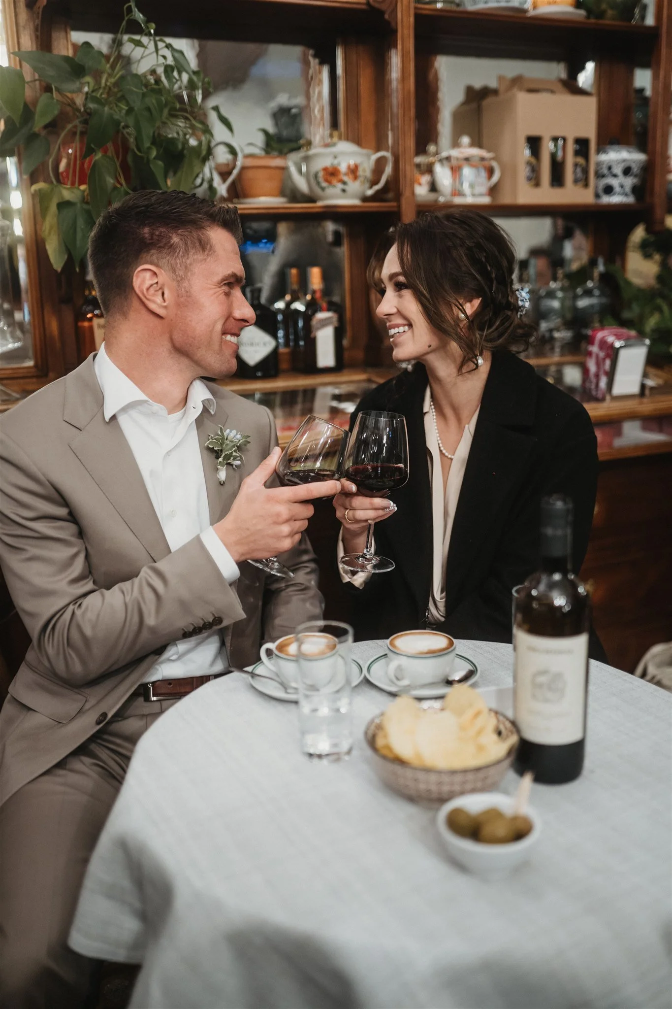 A couple dressed in formal attire sitting at a table, clinking glasses of red wine, smiling at each other in a cozy, vintage-style restaurant with wooden shelves and decorative teapots in the background. The table has coffee cups, a bread basket, and