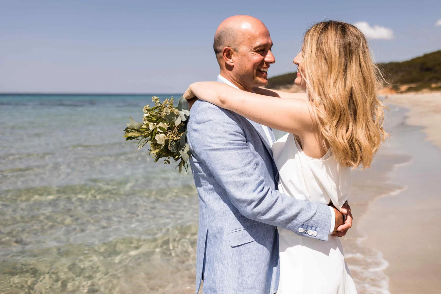 A man and woman embrace on a beach, smiling at each other, with the ocean and sky in the background.
