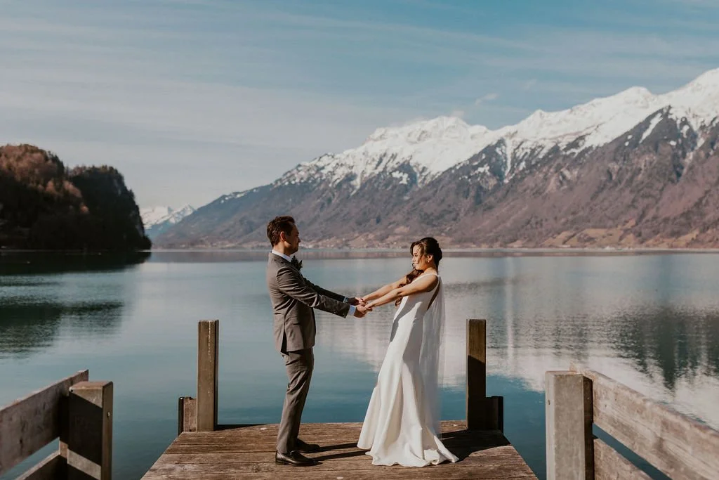 A bride and groom holding hands on a wooden dock by a lake with snow-capped mountains in the background.