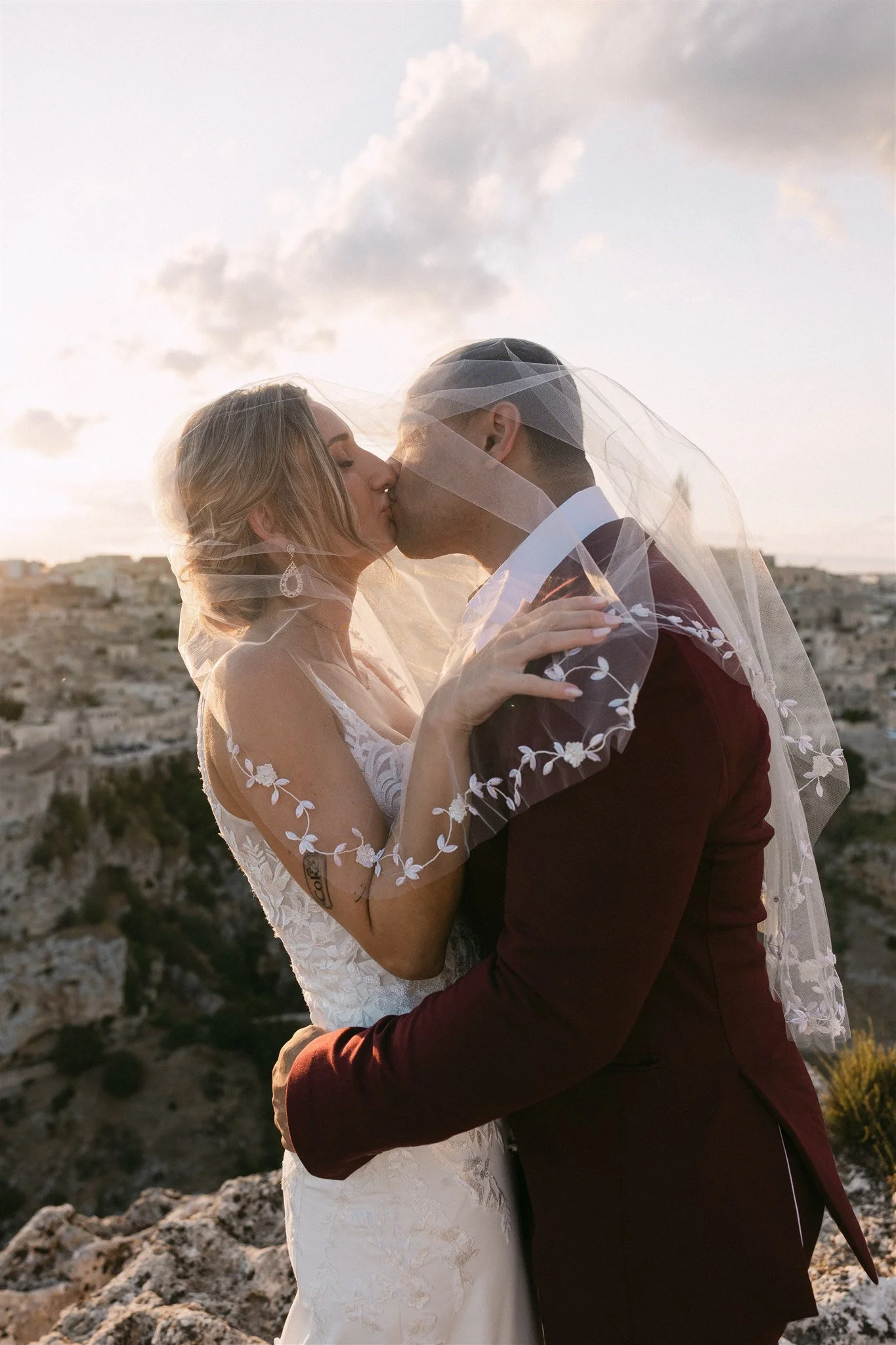 A bride and groom kissing outdoors at sunset, with the bride wearing a white dress and veil, and the groom in a dark suit, on a rocky landscape with a city in the background.