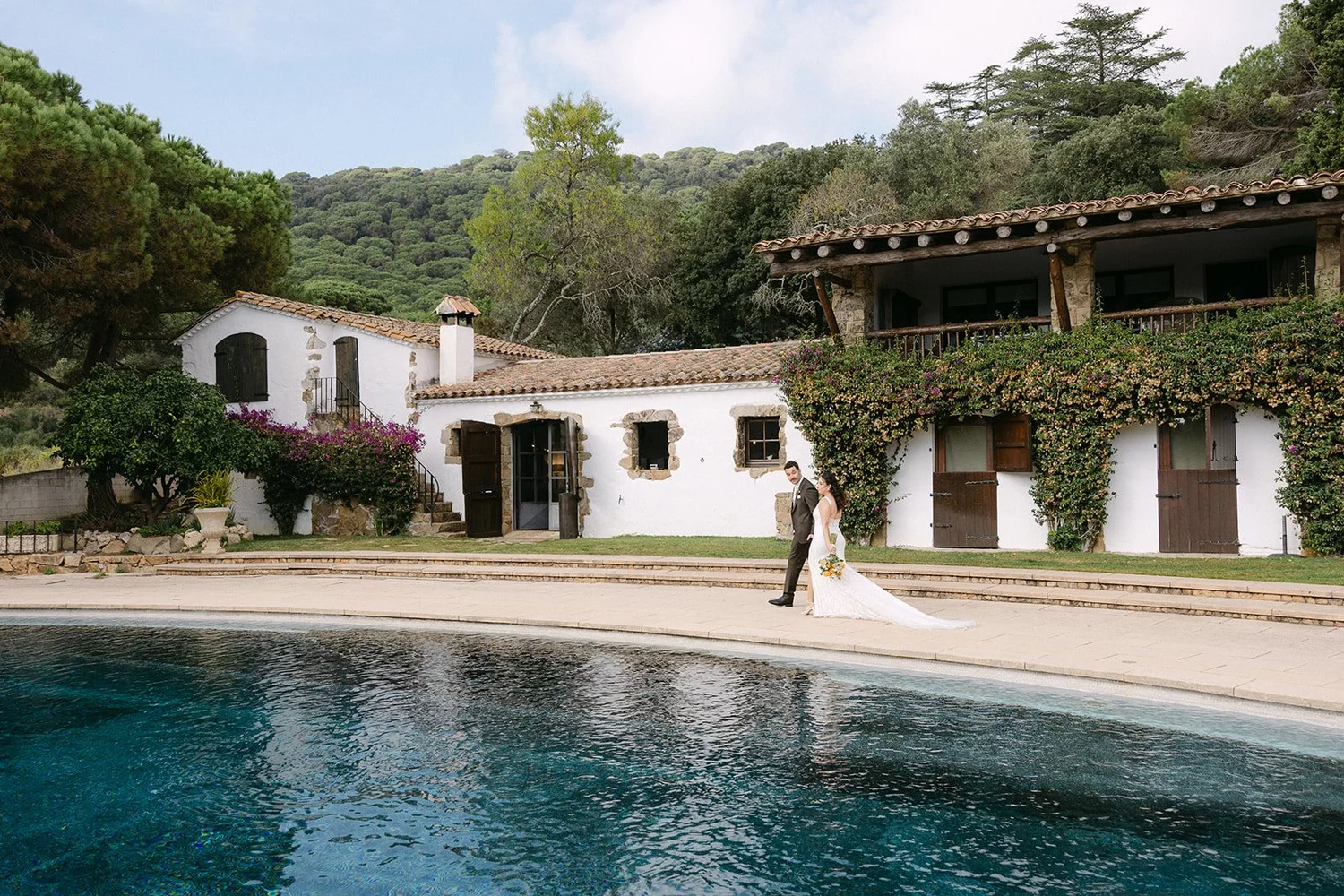 A bride and groom walking by a swimming pool in front of a rustic, white stucco house with wooden doors and windows, surrounded by greenery and colorful flowers, with hills in the background.