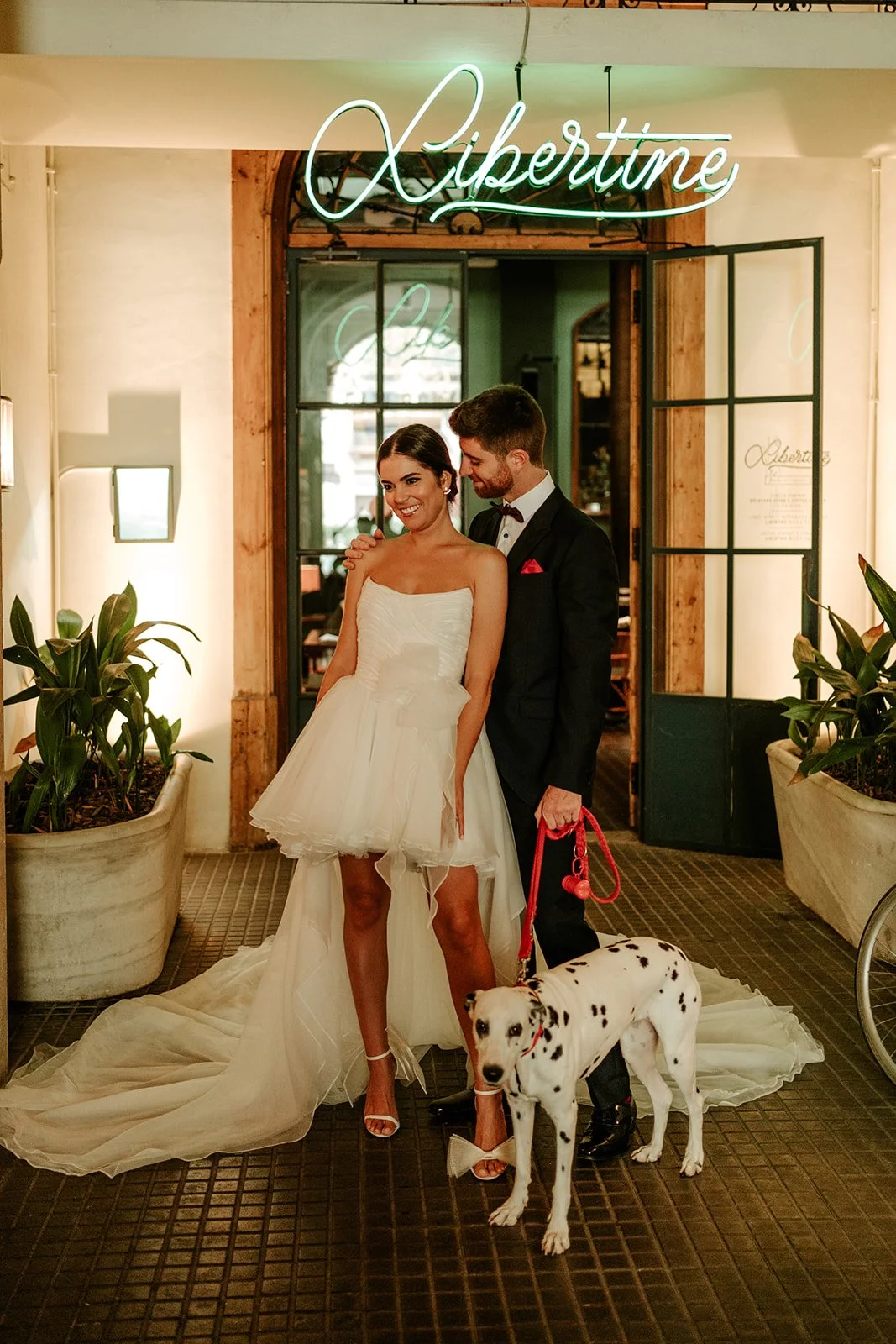 A newlywed couple stands indoors, the bride in a strapless white wedding dress and the groom in a black tuxedo. They are smiling with the groom holding a leash attached to a Dalmatian dog. A neon sign above reads "Libertine" and plants are on either side.