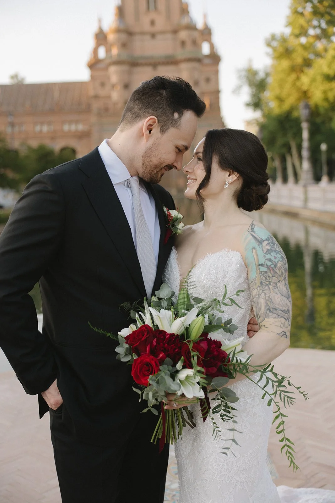 A newlywed couple in wedding attire standing outdoors by a body of water with a historic brick building in the background. The bride is holding a bouquet of red and white flowers, and they are smiling with their foreheads touching.