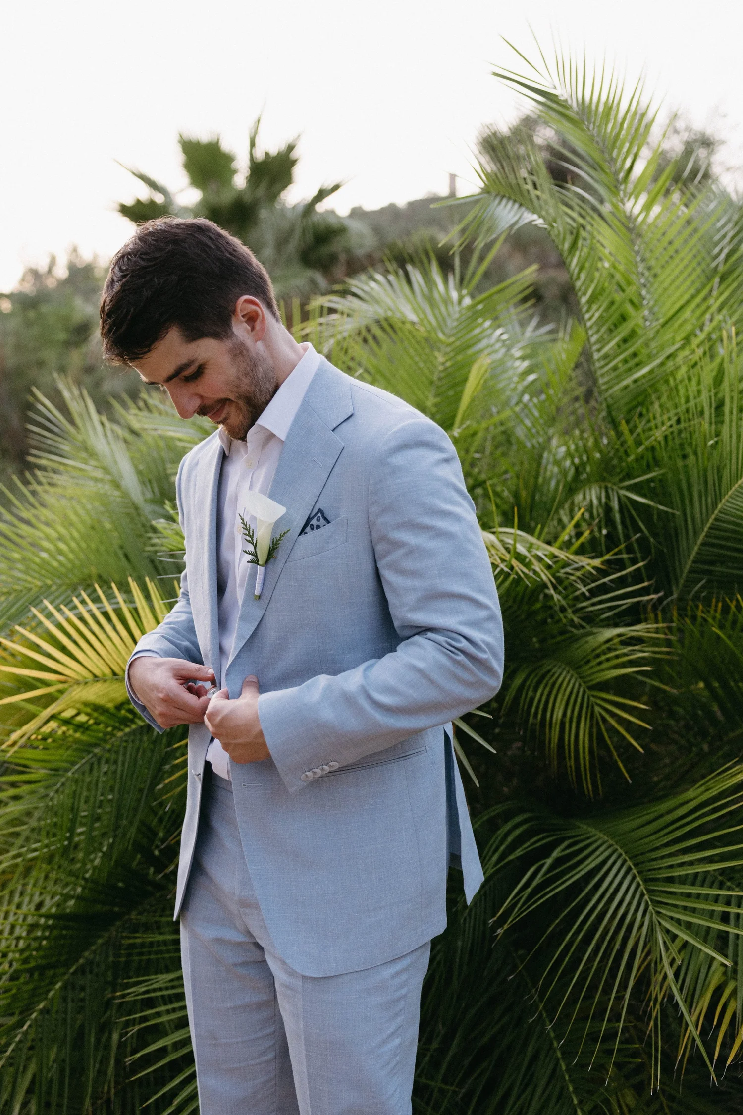 A man in a light gray suit with a white dress shirt and boutonniere, standing outdoors among green palm leaves, looking down and buttoning his suit jacket.