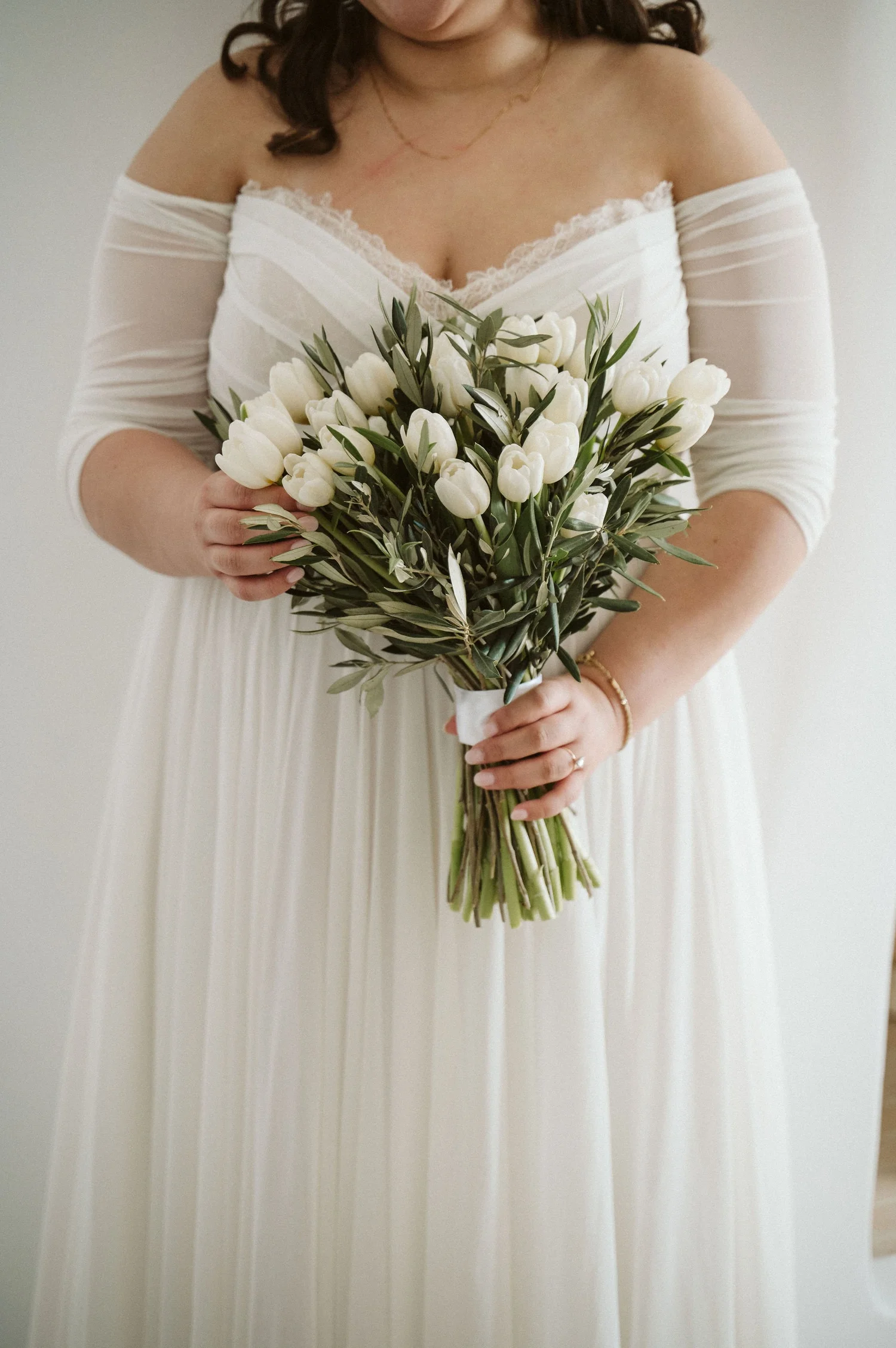 A woman in an off-the-shoulder white dress with lace detailing, holding a bouquet of white tulips with green leaves, standing against a plain light background.
