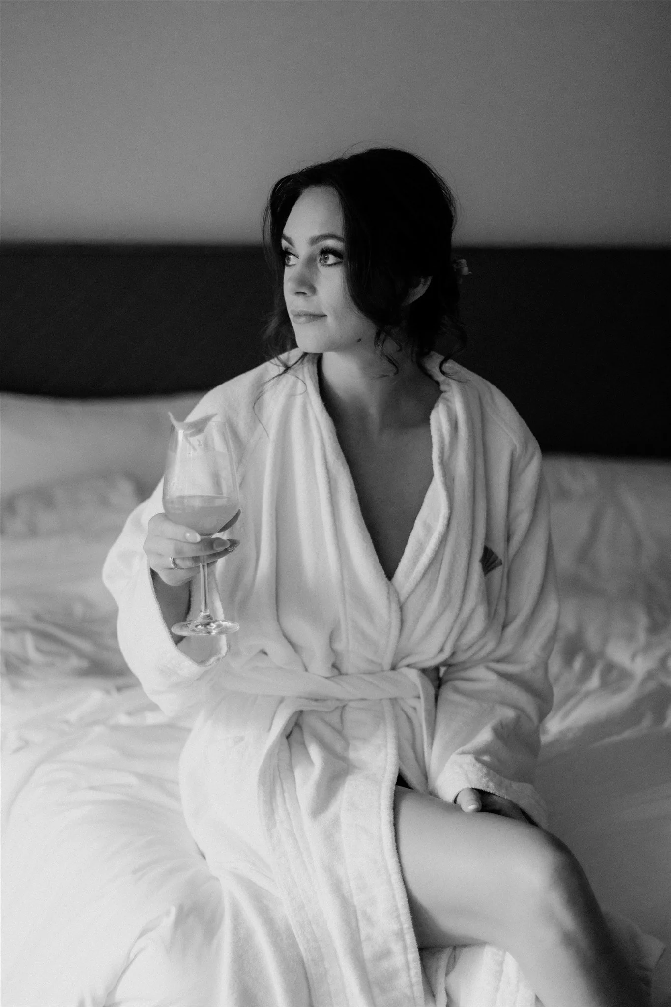A woman with dark hair sitting on a bed, wearing a white robe, holding a glass of wine, looking to the side, in a relaxed setting.