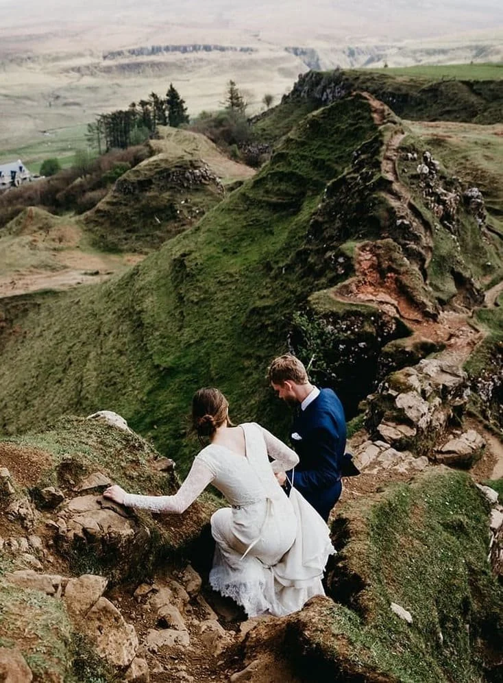A bride and groom in wedding attire climbing a rocky, grassy hillside with scenic green valleys and cliffs in the background.