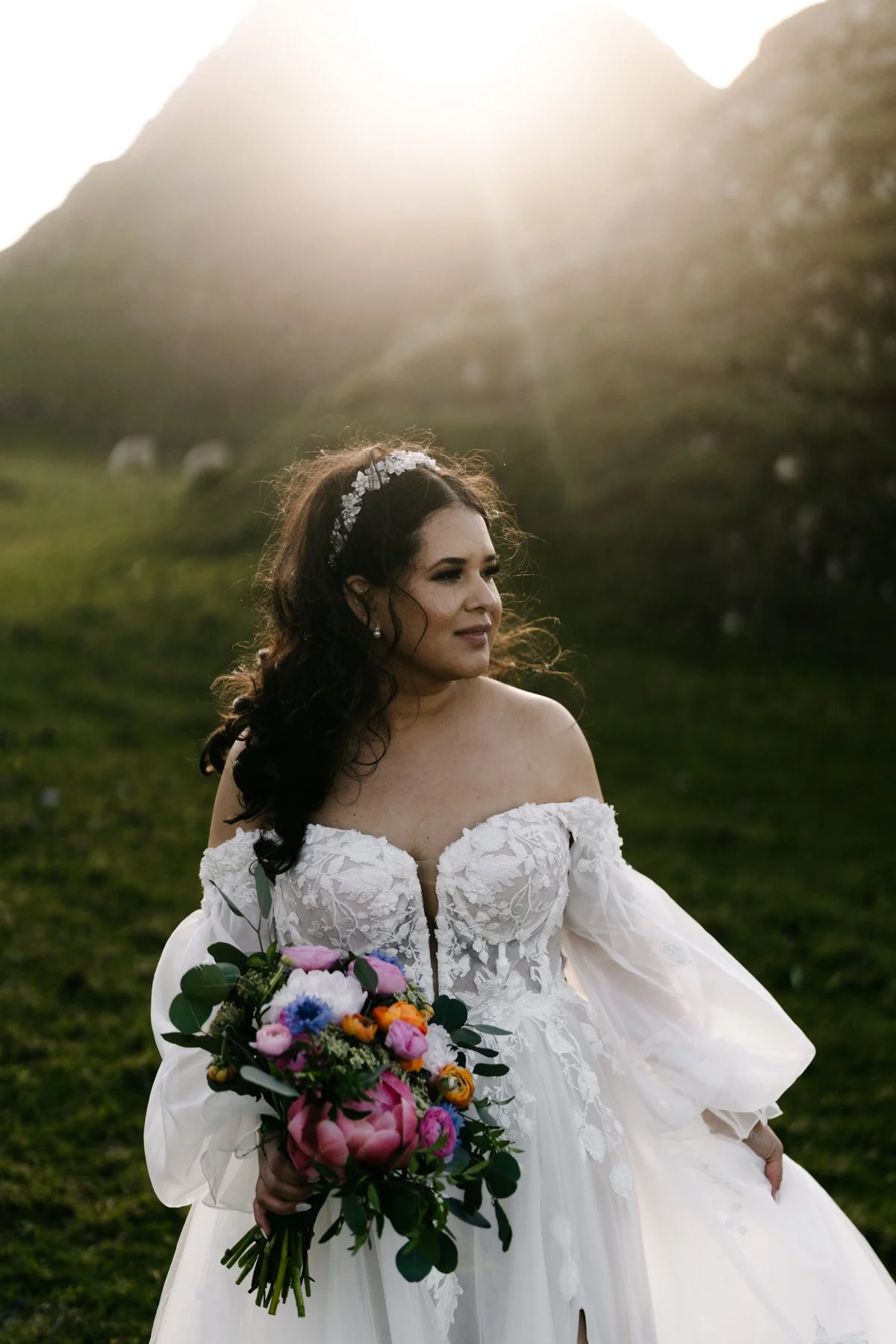 Elopement portrait of a woman in a wedding dress holding a colorful bouquet of flowers outdoors during sunset, with mountains in the background.