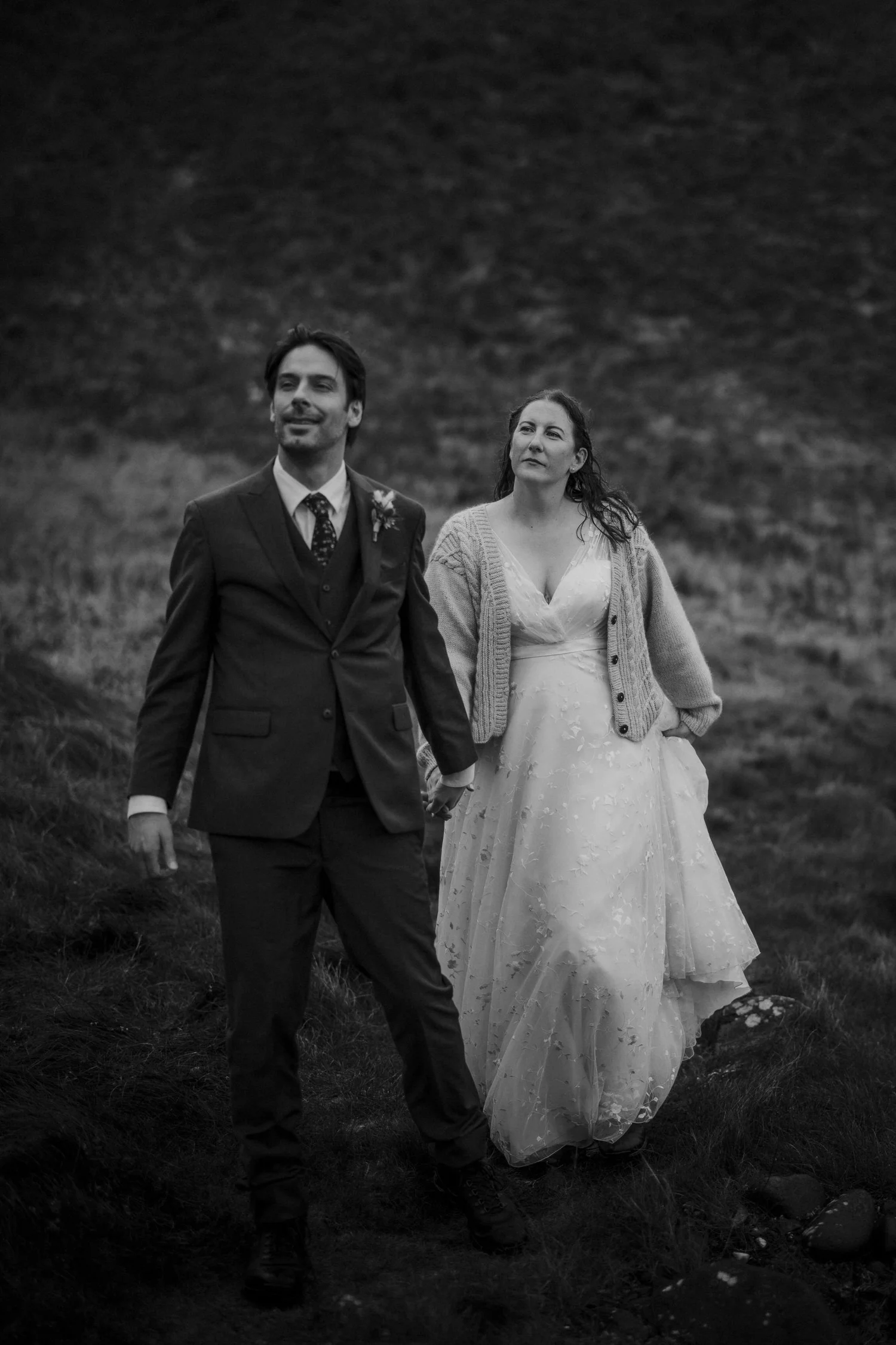 A black and white photo of a man in a suit and a woman in a wedding dress walking outdoors on grassy terrain.