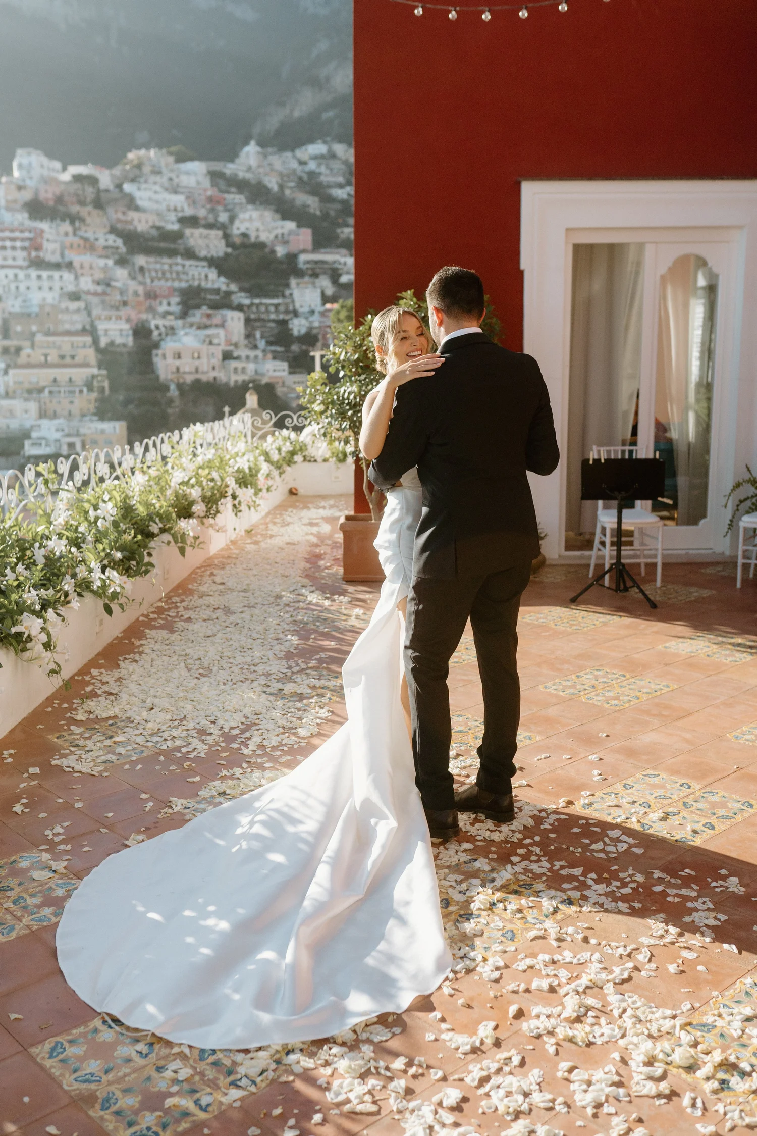 A bride and groom dancing on a terrace during their wedding, with white flower petals scattered on the floor and a scenic hillside town in the background.