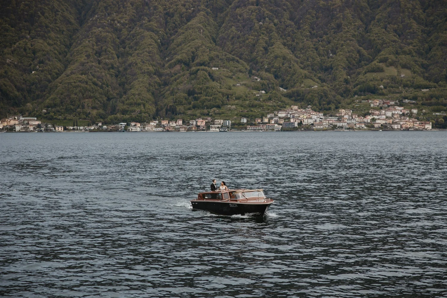 A small boat with two people on board is sailing on a large body of water with a town and green, forested hills in the background.