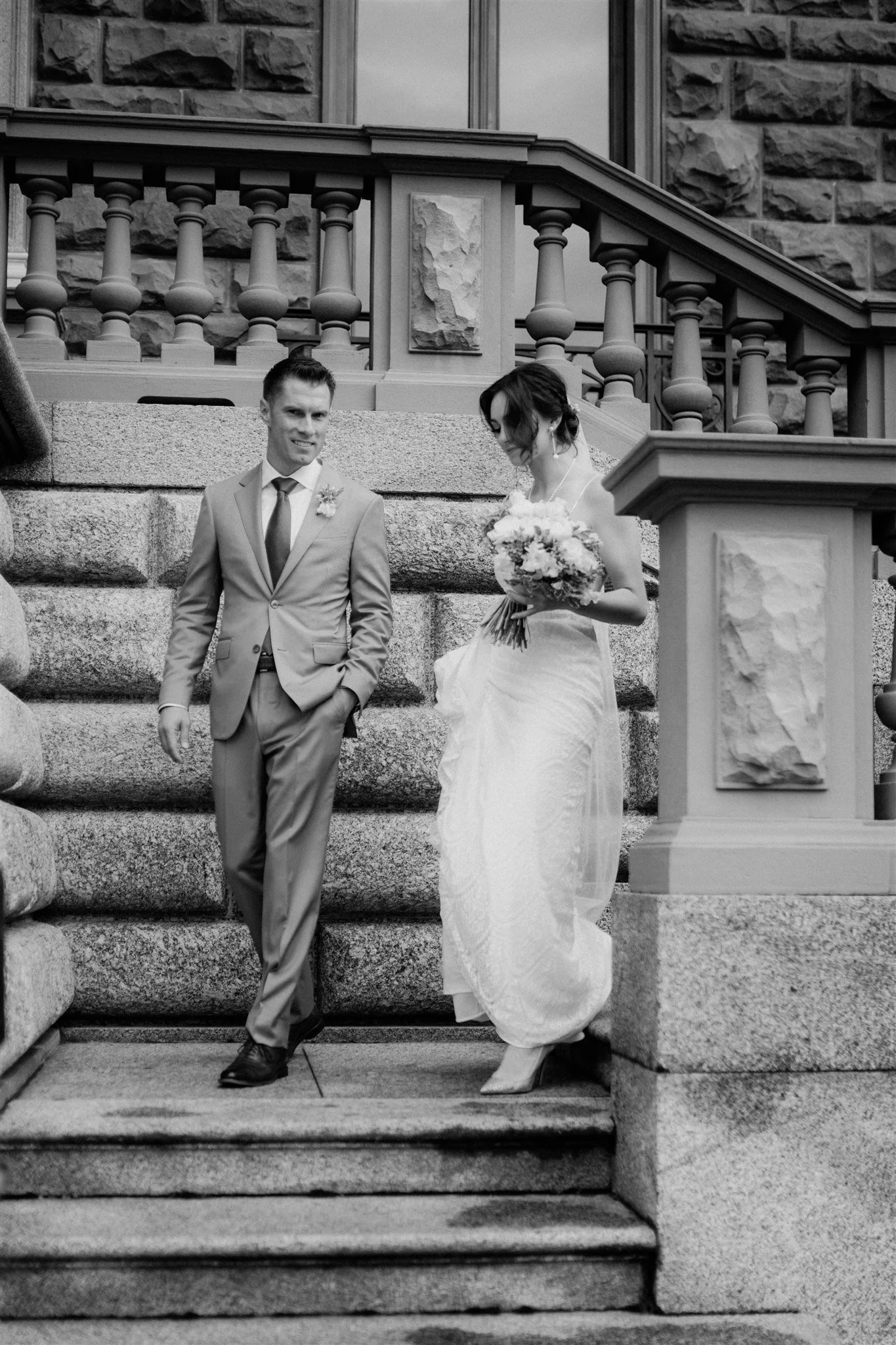 A black-and-white photo of a bride and groom standing on outdoor stone steps near a stone building, with the groom smiling and the bride looking down at her bouquet.
