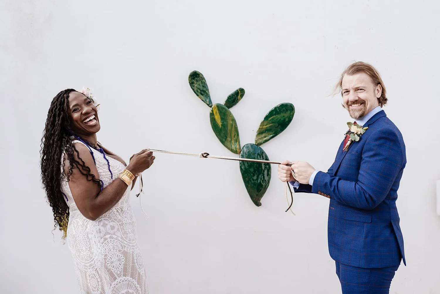 A couple dressed in wedding attire holding a rope, smiling, with a white wall and a green cactus decoration in the background.