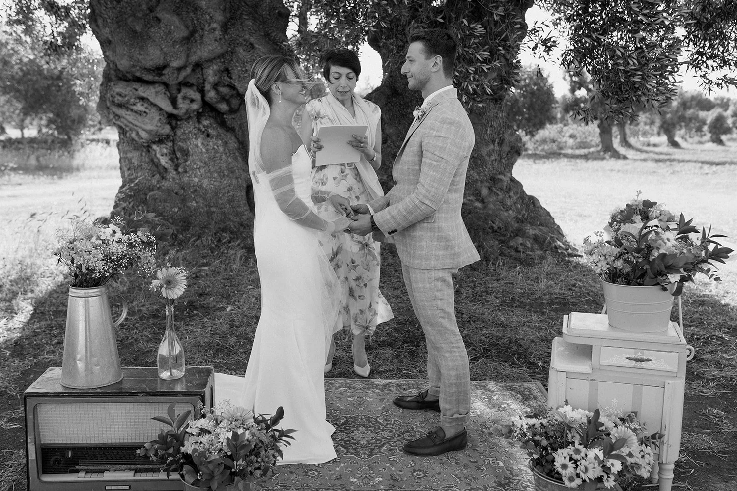 A black and white photograph of a wedding ceremony outdoors under a large tree. The bride and groom are holding hands, facing each other, with a woman reading vows or giving a speech. Flowers and vintage decor are arranged around them.