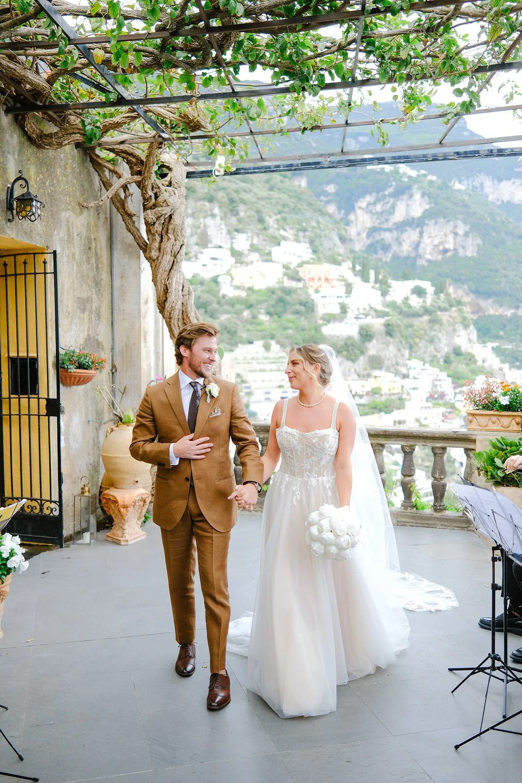 A bride and groom holding hands and smiling during their wedding ceremony outdoors on a terrace with scenic mountain views. The bride is wearing a white wedding dress and holding a bouquet, while the groom is dressed in a brown suit with a boutonnièr