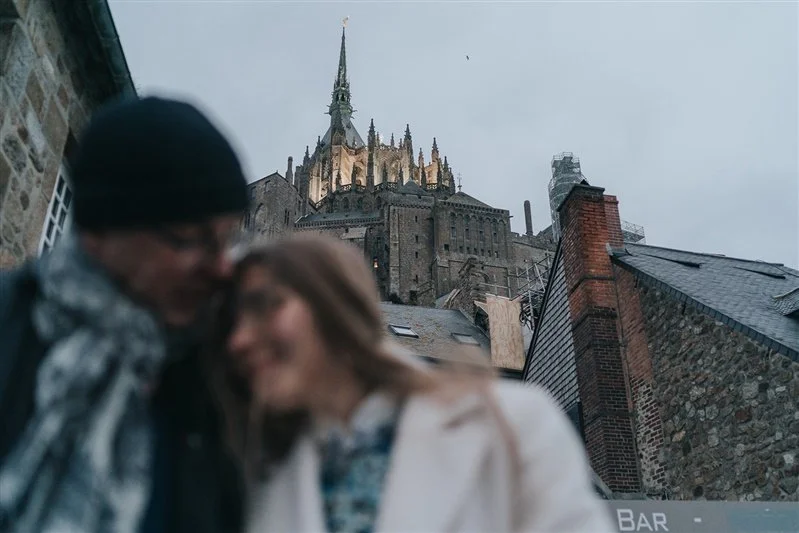 Blurred couple smiling and leaning into each other in front of a historic castle or church with a tall spire, brick and stone buildings, and cloudy sky.