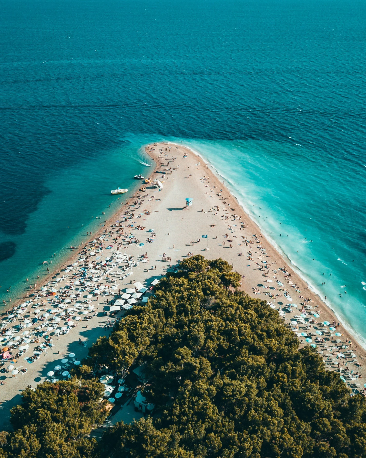 An aerial view of a sandy beach with many umbrellas and people, bordered by lush green trees, and extending into the blue ocean with a small boat near the shore.