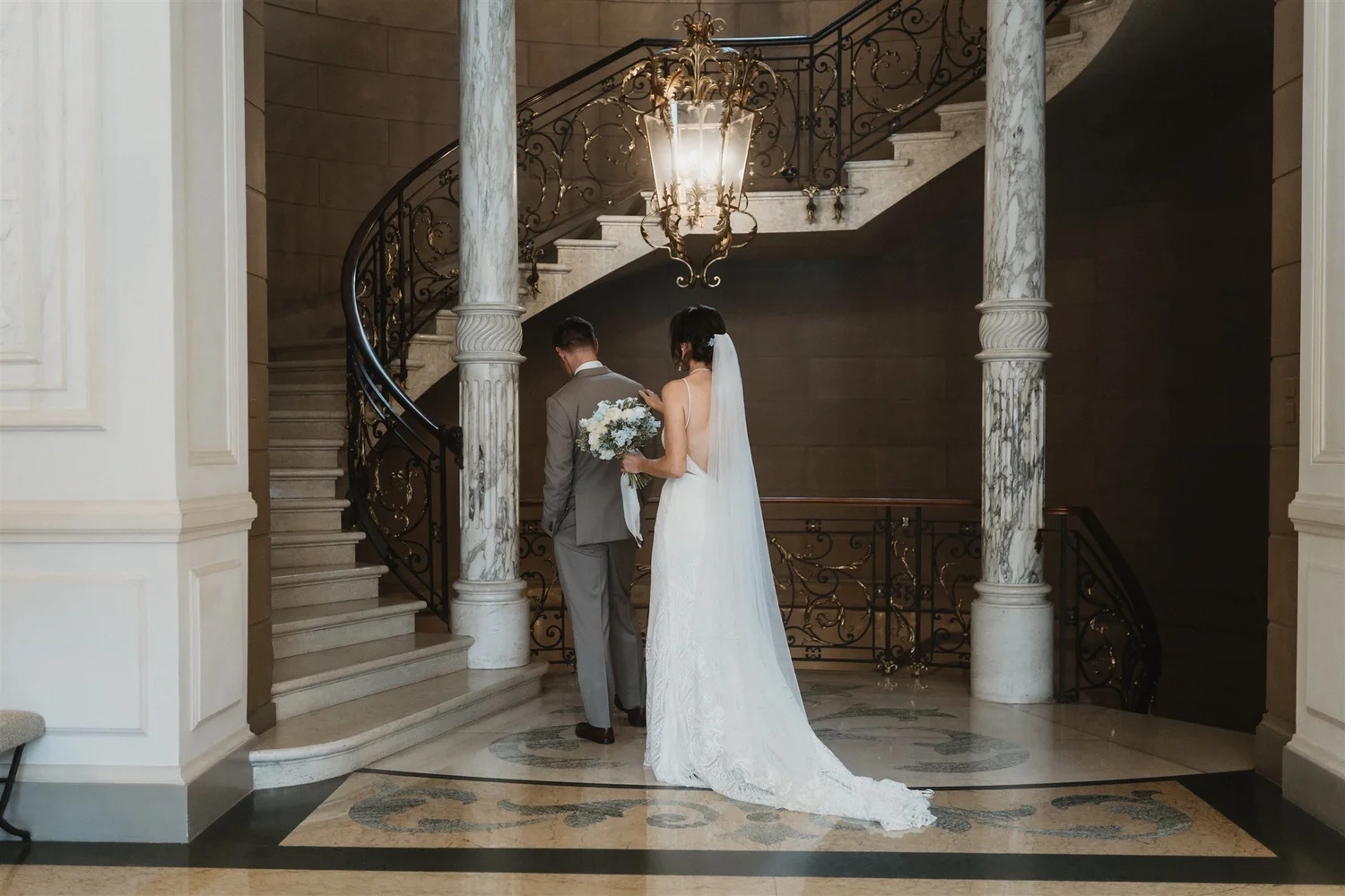 A bride and groom stand at the bottom of a grand staircase inside a elegant building. The bride is wearing a white wedding dress and veil, holding a bouquet of white flowers. The groom is wearing a light gray suit. A large ornate chandelier hangs abo